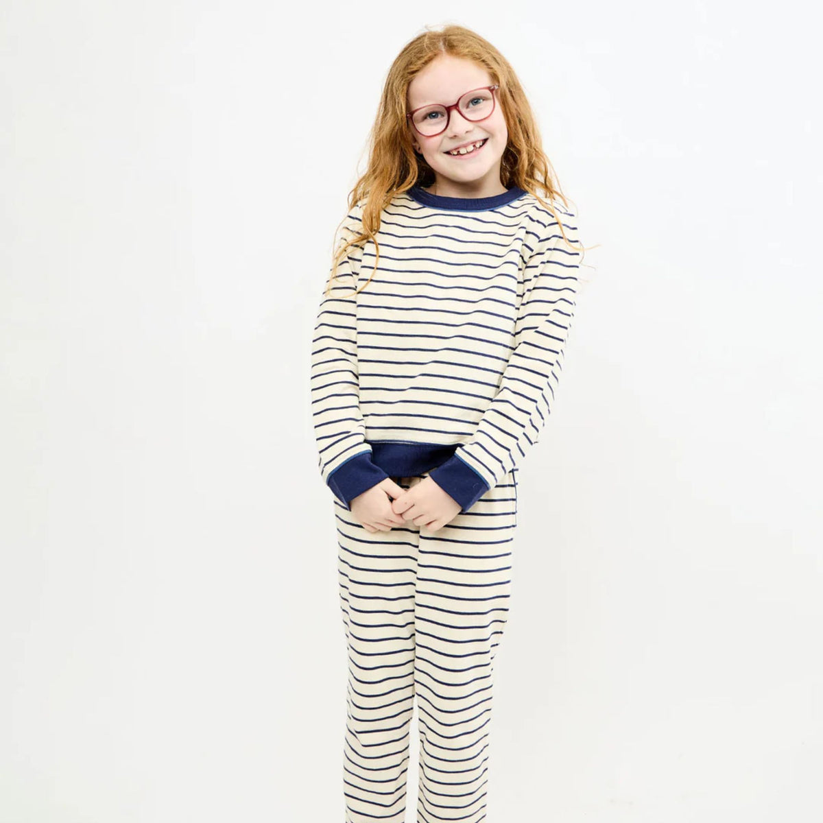 A young girl with glasses wearing a matching two-piece outfit with navy blue and cream horizontal stripes, standing against a plain white background.