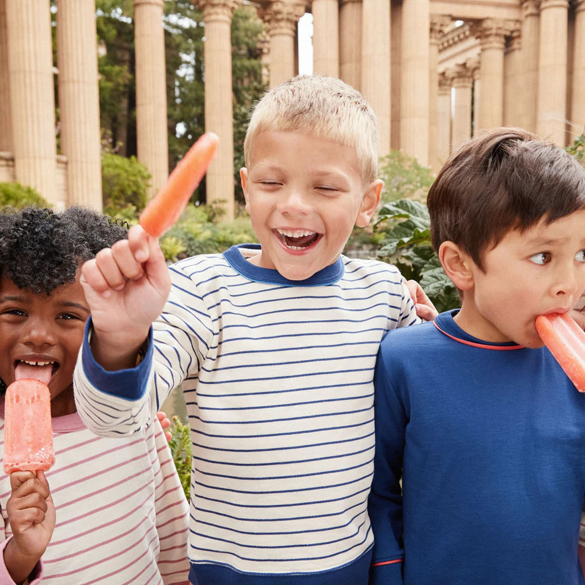Three children outdoors in front of large stone columns, each holding or biting a pink popsicle. The child in the center wears a white long-sleeve shirt with thin navy stripes, while the child on the right wears a solid navy shirt.