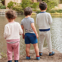 Three young children standing on a shore by a lake, viewed from behind. They are wearing striped long-sleeve shirts and matching pants or shorts in soft pastel and navy tones.