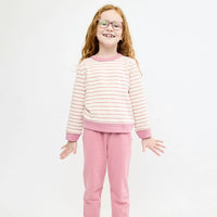 A young girl with glasses and wavy light brown hair stands against a plain white studio background, wearing a two-piece outfit consisting of a cream and pink striped long-sleeve top with solid pink trim, and matching solid pink pants.