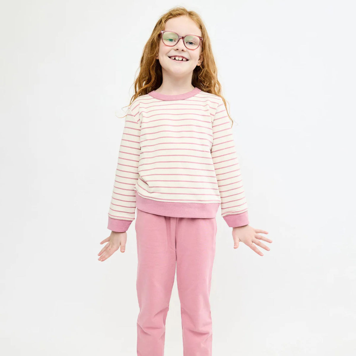 A young girl with glasses and wavy light brown hair stands against a plain white studio background, wearing a two-piece outfit consisting of a cream and pink striped long-sleeve top with solid pink trim, and matching solid pink pants.