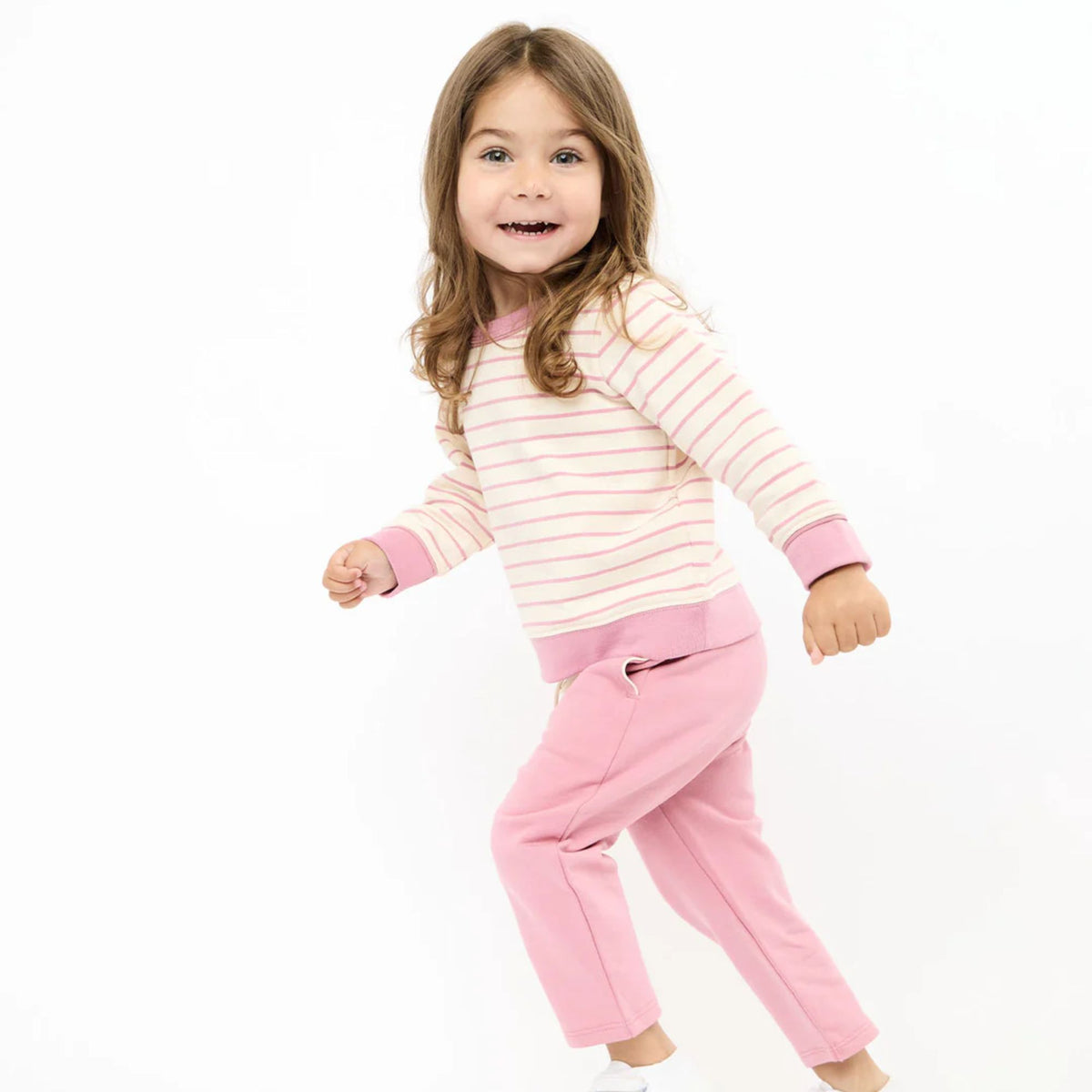 A young girl with long brown hair is wearing a two-piece outfit consisting of a light pink and white striped long-sleeve top with solid pink cuffs and collar, paired with solid pink pants, against a plain white background.