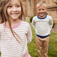 Two children standing outdoors on grass in front of a fence.  The child in the foreground wears a pink and white striped long-sleeve shirt, and the child in the background wears a white long-sleeve shirt with thin navy horizontal stripes and tan pants.