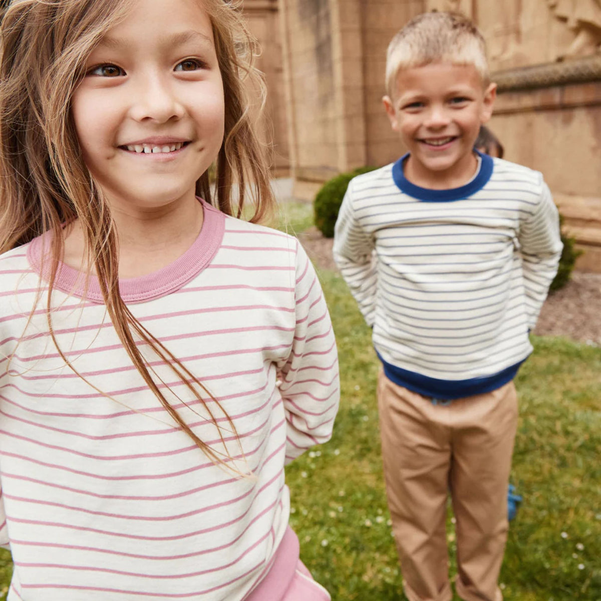 Two children standing outdoors on grass in front of a fence.  The child in the foreground wears a pink and white striped long-sleeve shirt, and the child in the background wears a white long-sleeve shirt with thin navy horizontal stripes and tan pants.