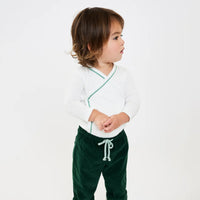 A toddler standing against a plain white studio background, wearing a white long-sleeve wrap-style top with thin mint green piping and dark green drawstring corduroy pants.