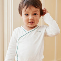 A baby wearing a white long-sleeve wrap onesie with an off to the side light blue-green trim and standing against a soft beige paneled background.