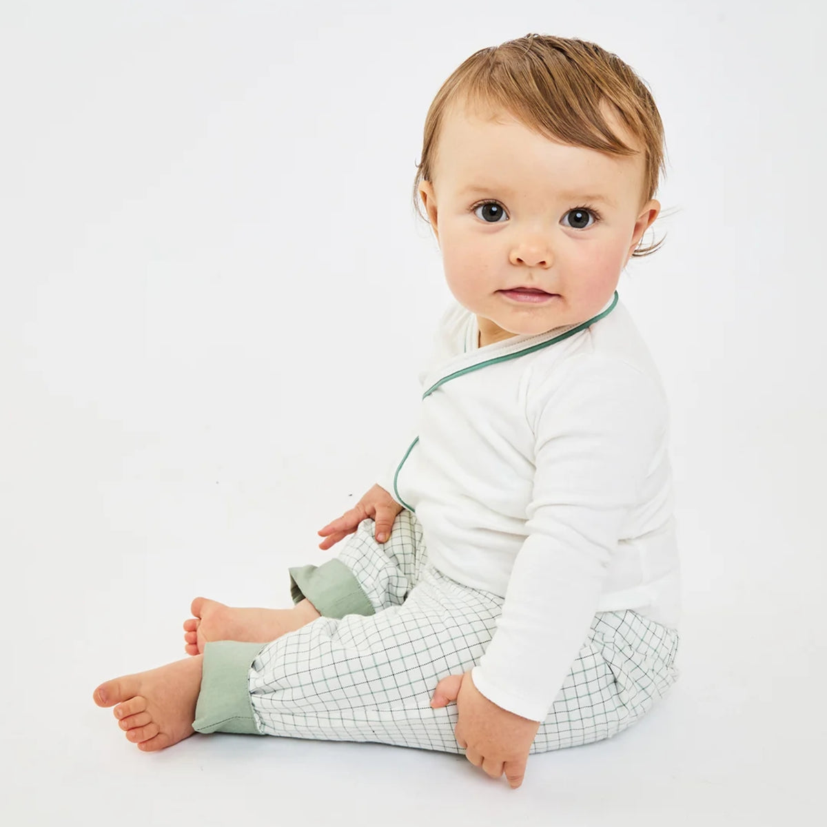 A baby sitting on a floor wearing a white long-sleeve top with green trim and green-and-white checkered pants.