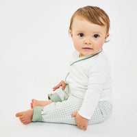 A baby sitting on a floor wearing a white long-sleeve top with green trim and green-and-white checkered pants.