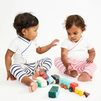 Two toddlers sitting on a white floor playing with colorful wooden toy trains, wearing white wrap-style baby outfits. One outfit has navy stripes, and the other has pink stripes.