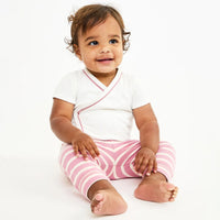 A baby sitting on a white background wearing a white short-sleeve wrap-style top with pink trim and pink-and-white striped pants.