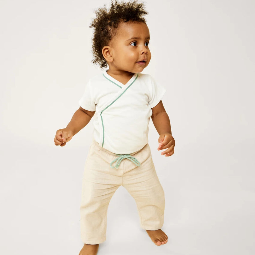 A toddler standing against a light gray  background, wearing a white short-sleeve wrap-style top with mint green trim and cream pants with a mint green drawstring.