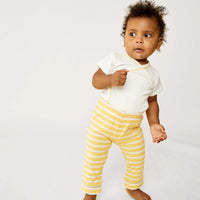 A toddler standing barefoot against a plain light background, wearing a white short-sleeve top with yellow trim and yellow-and-white striped pants.