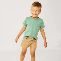 A young boy modeling a light green short-sleeve pocket t-shirt with blue trim and tan shorts against a plain white studio background.
