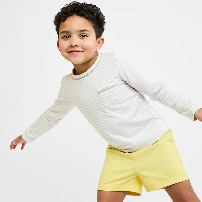 A young boy wearing a long-sleeve white shirt with thin gray stripes and yellow shorts, posing against a clean white studio background.