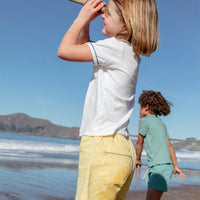 Two children on a beach with one wearing a white shirt and yellow shorts, and the other in a green shirt.