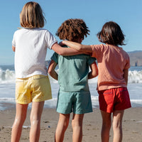 Three children standing on a beach with their backs to the camera, wearing colorful short-sleeve shirts and shorts in white, yellow, mint green, and coral pink.