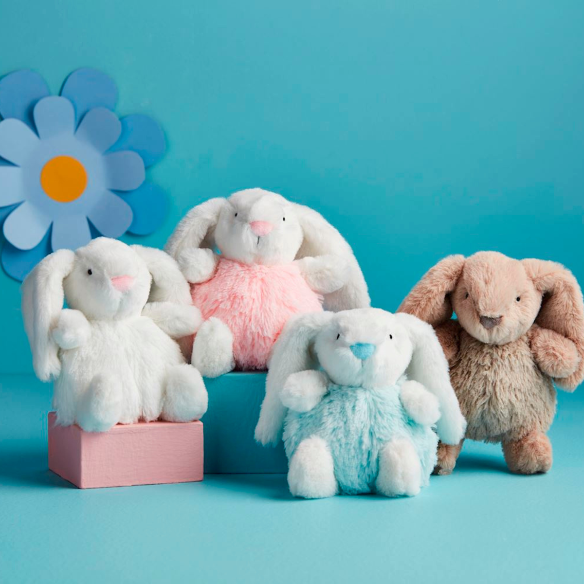 Four plush bunny toys in different colors on a blue background with a large flower decoration.