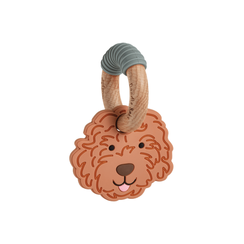 A baby teething toy featuring a sculpted golden doodle dog face in a warm terracotta-brown color, attached to a natural wood ring with a gray ribbed silicone loop at the top, shown on a plain white background.