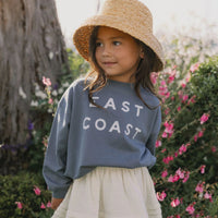 A young girl wearing a straw bucket hat, a slate blue sweatshirt with 'EAST COAST' printed in white, and a cream skirt, standing outdoors in front of a tree and pink flowers.