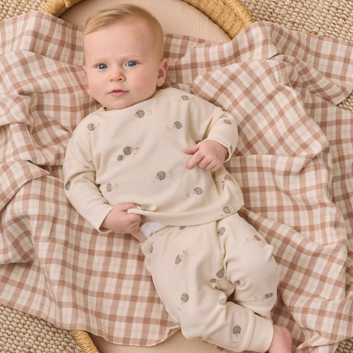 A baby lying on a woven blanket, wearing a cream long-sleeve pajama set with small gray and brown woodland animal prints. 