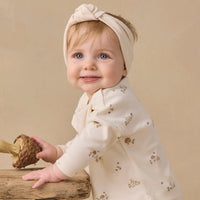 A baby wearing a cream-colored long-sleeve romper with a small brown mushroom pattern and a matching knotted headband, posed on a rustic wooden surface against a warm beige background.