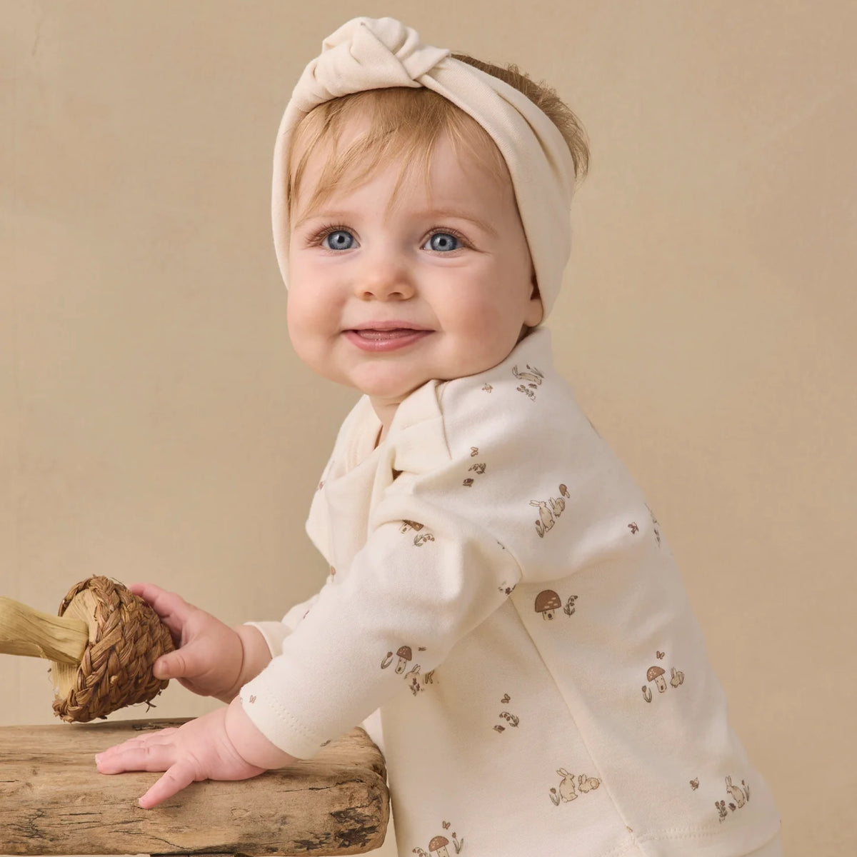 A baby wearing a cream-colored long-sleeve romper with a small brown mushroom pattern and a matching knotted headband, posed on a rustic wooden surface against a warm beige background.