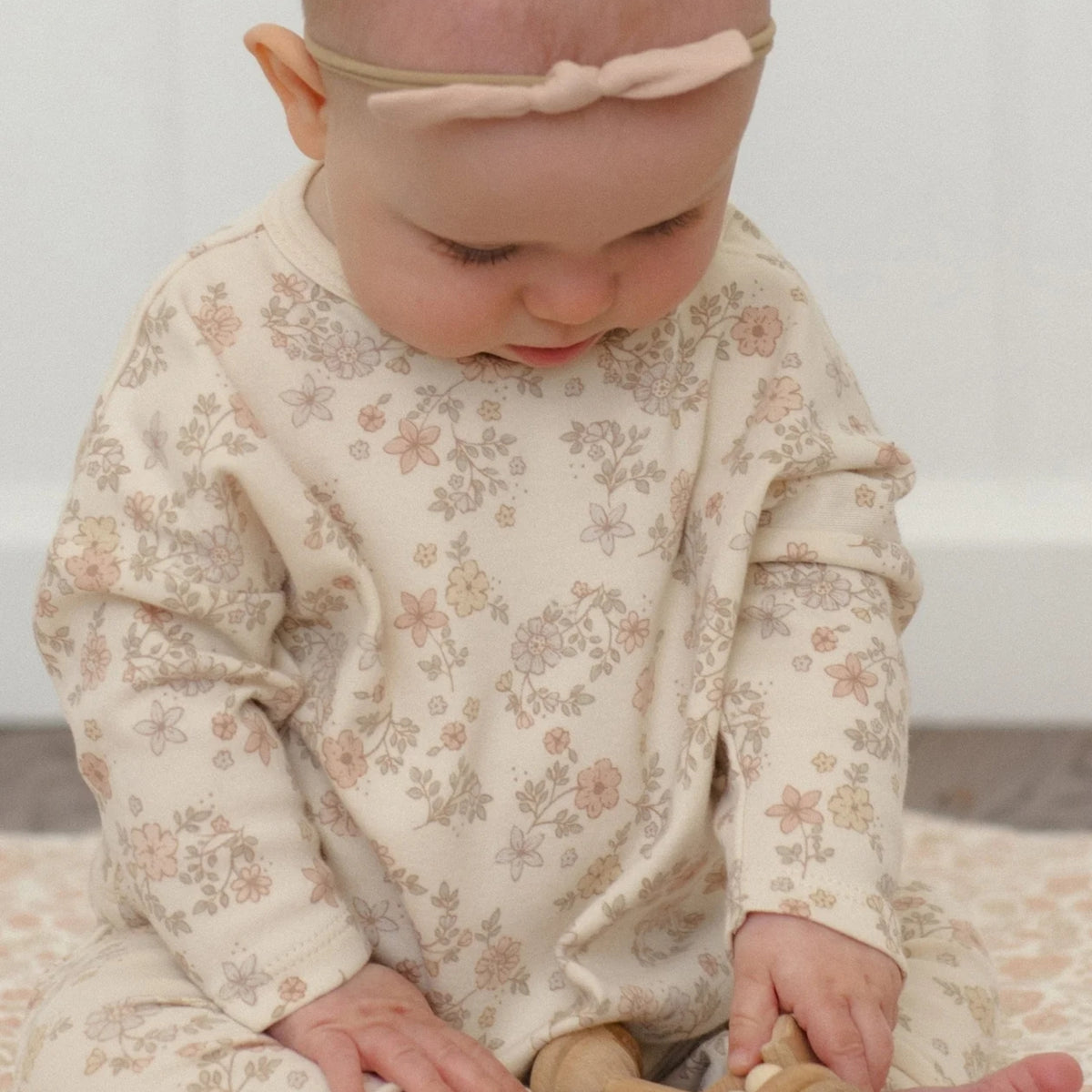 A baby wearing a cream-colored long-sleeve footless romper with a delicate pink and beige floral pattern, sitting on a patterned rug.