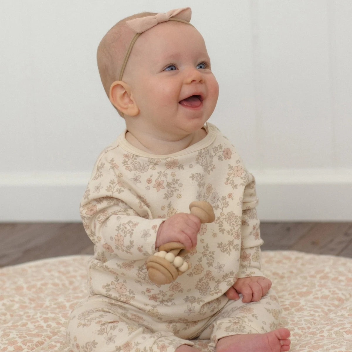 A baby wearing a cream floral long-sleeve jumpsuit and a matching bow headband, sitting on a patterned rug and holding a wooden rattle.