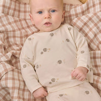 A baby lying on a beige and brown gingham blanket, wearing a cream-colored long-sleeve romper with small brown and gold hedgehog prints.