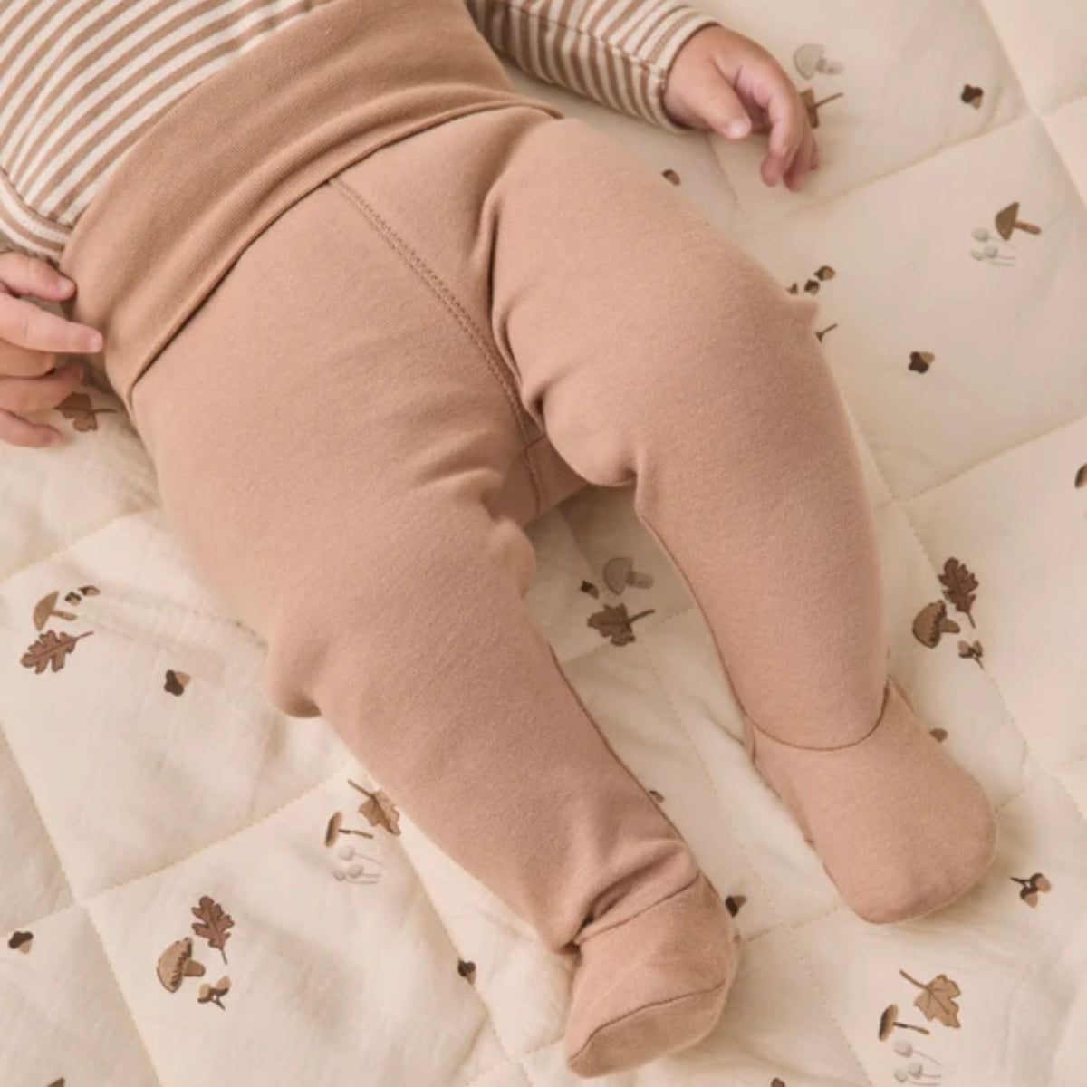A close-up of a baby lying on a quilted cream blanket with a small brown leaf print, wearing solid tan fleece footed leggings and a matching striped top.