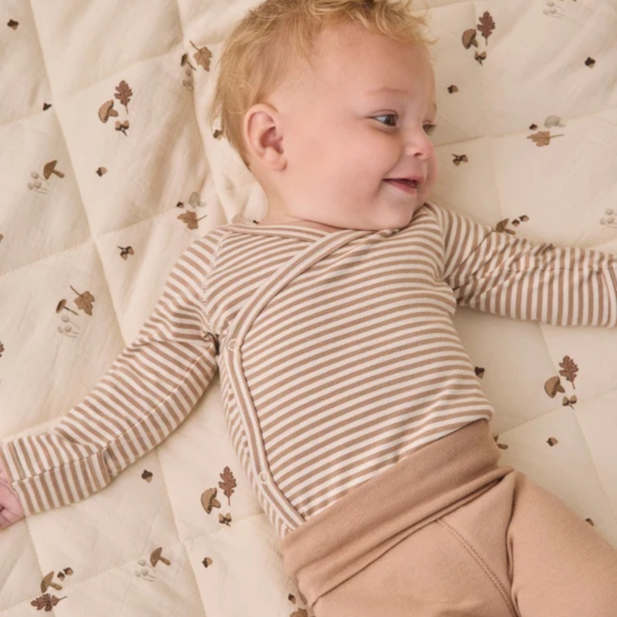 A baby lying on a quilted cream mattress  wearing a beige and white striped long-sleeve wrap bodysuit and tan pants.