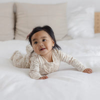 A baby wearing a cream and beige patterned long-sleeve romper lies on a white bed, looking up and smiling.