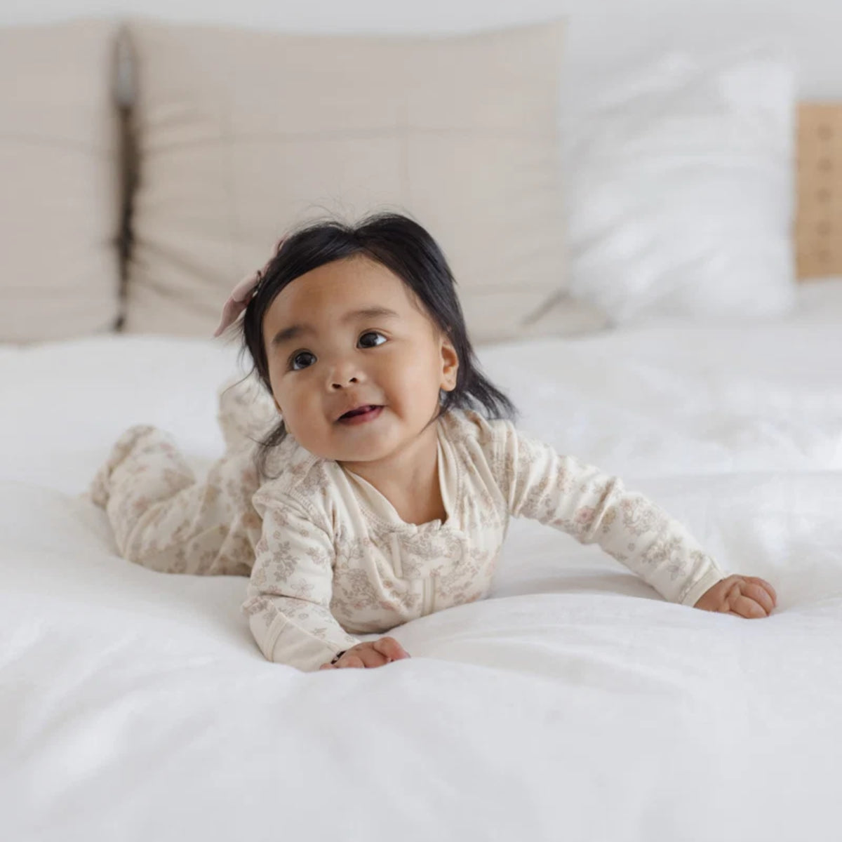 A baby wearing a cream and beige patterned long-sleeve romper lies on a white bed, looking up and smiling.