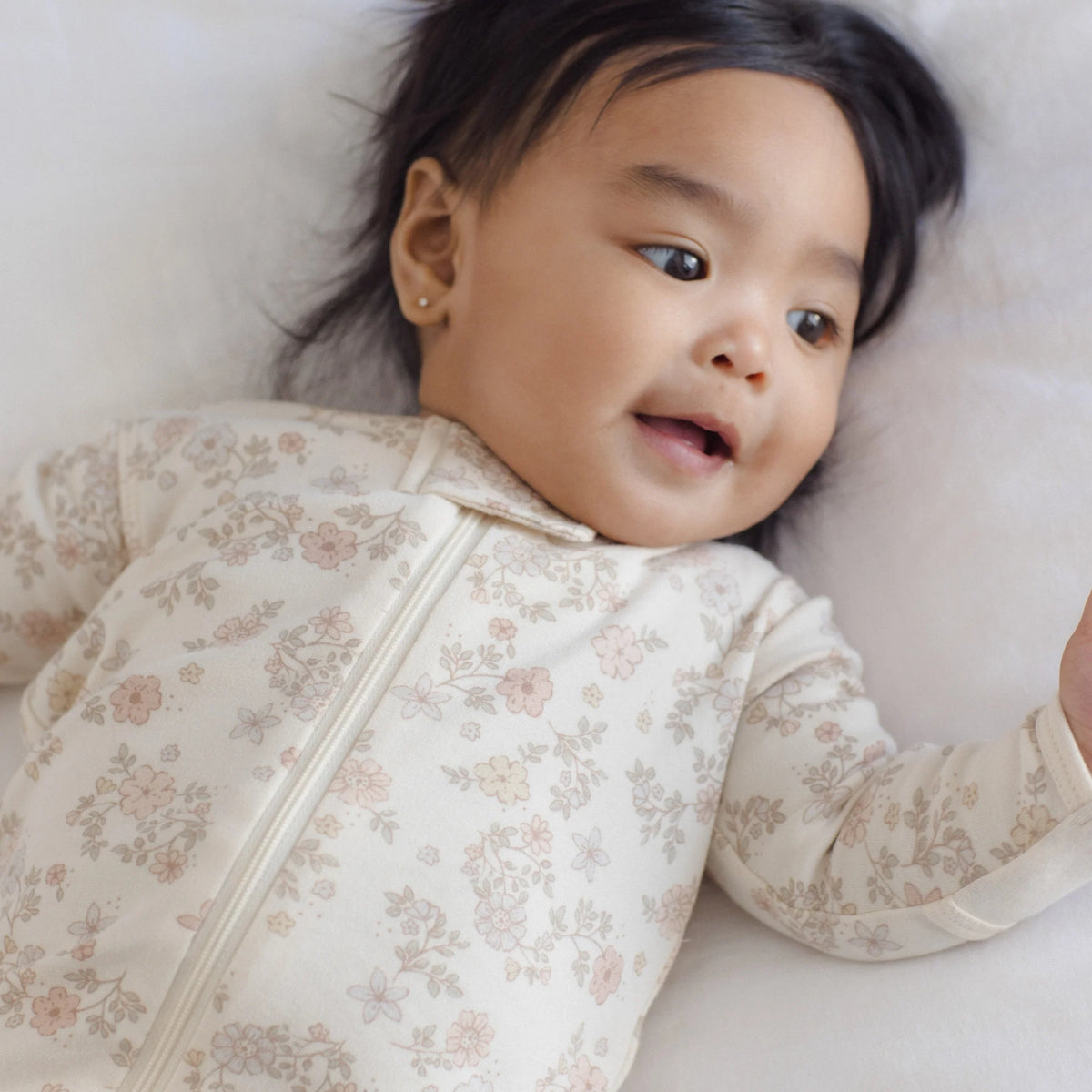 A baby lying on a soft white surface, wearing a long-sleeve cream-colored zip-up sleeper with a delicate pink and beige floral pattern.