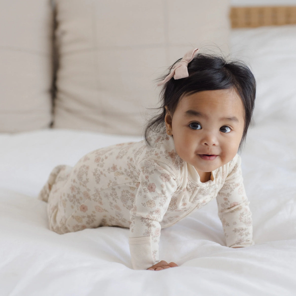 A baby crawling on a white bed wearing a cream-colored long-sleeve pajama with a delicate pink floral pattern and a matching pink bow in her hair.