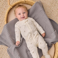 A baby lying on a gray textured blanket inside a woven basket, wearing an off-white footed sleeper with a small sheep print.