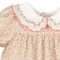 Close-up of a dress with a cream-colored collar featuring pink floral embroidery and a ruffled pink trim, paired with a light peach fabric printed with small green and pink flowers.