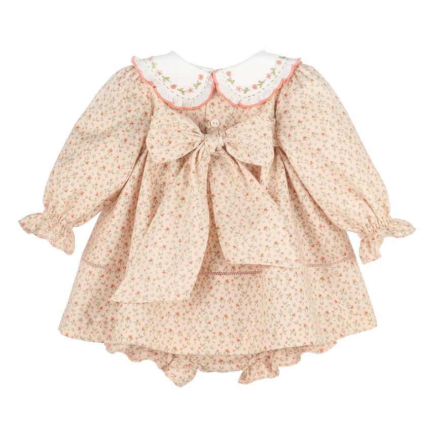 A girl's long-sleeve dress with a small floral print in beige, pink, and green, featuring a large bow at the center back, a white Peter Pan collar with pink trim, and ruffled cuffs and hem.