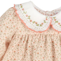Close-up of a dress with a cream Peter Pan collar featuring embroidered pink flowers and green leaves, surrounded by a pink ruffled trim, paired with a light peach floral bodice.