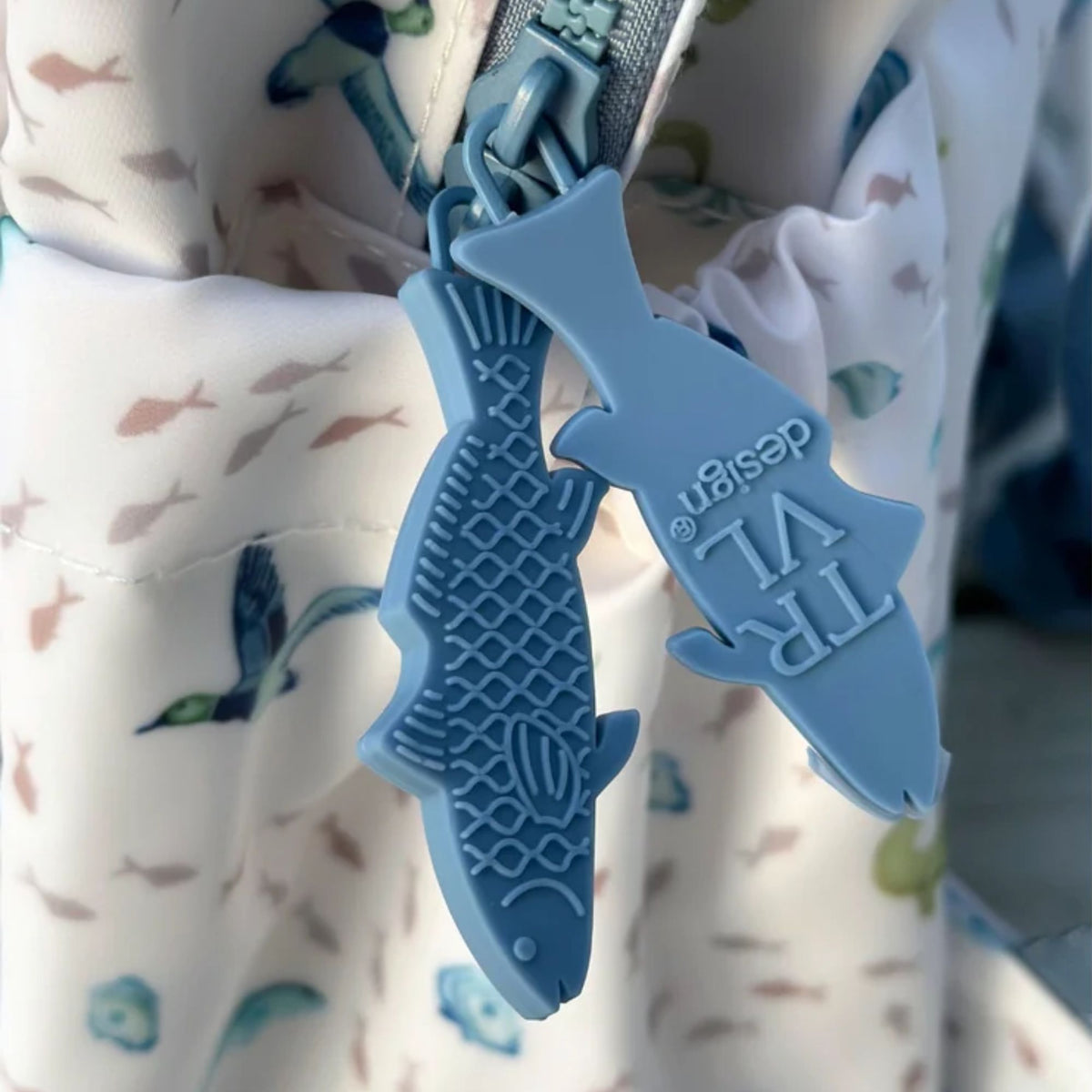 A close-up of a light blue zipper pull featuring two fish-shaped charms, one embossed with the text 'TRP VI design'. The zipper is set into a white fabric with a light blue and pink fish pattern.