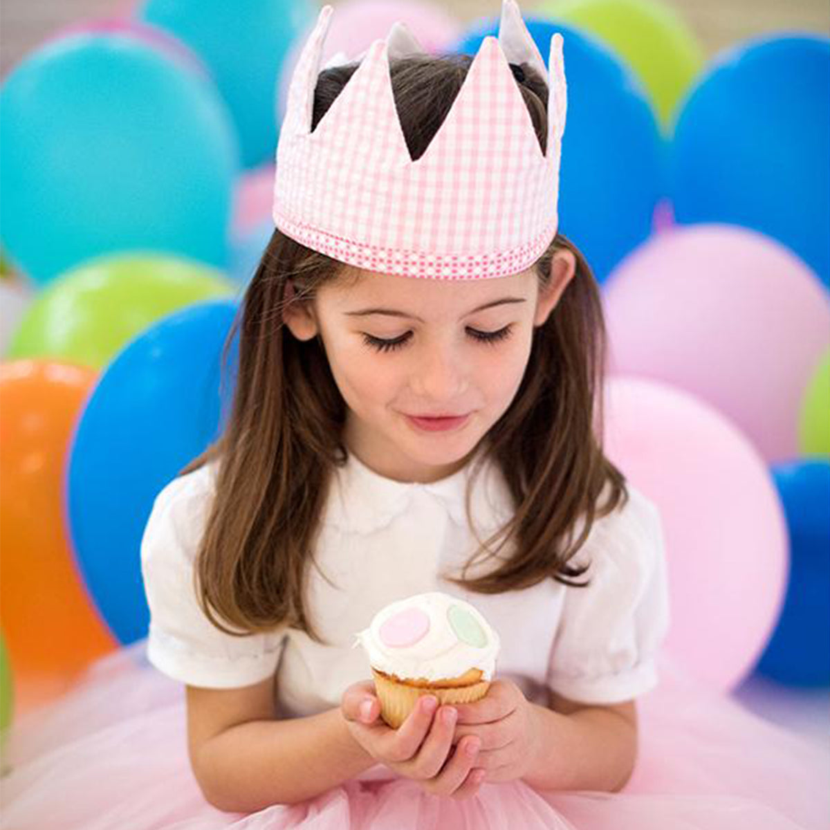 Pink gingham birthday crown atop the head of a girl holding a cupcake.